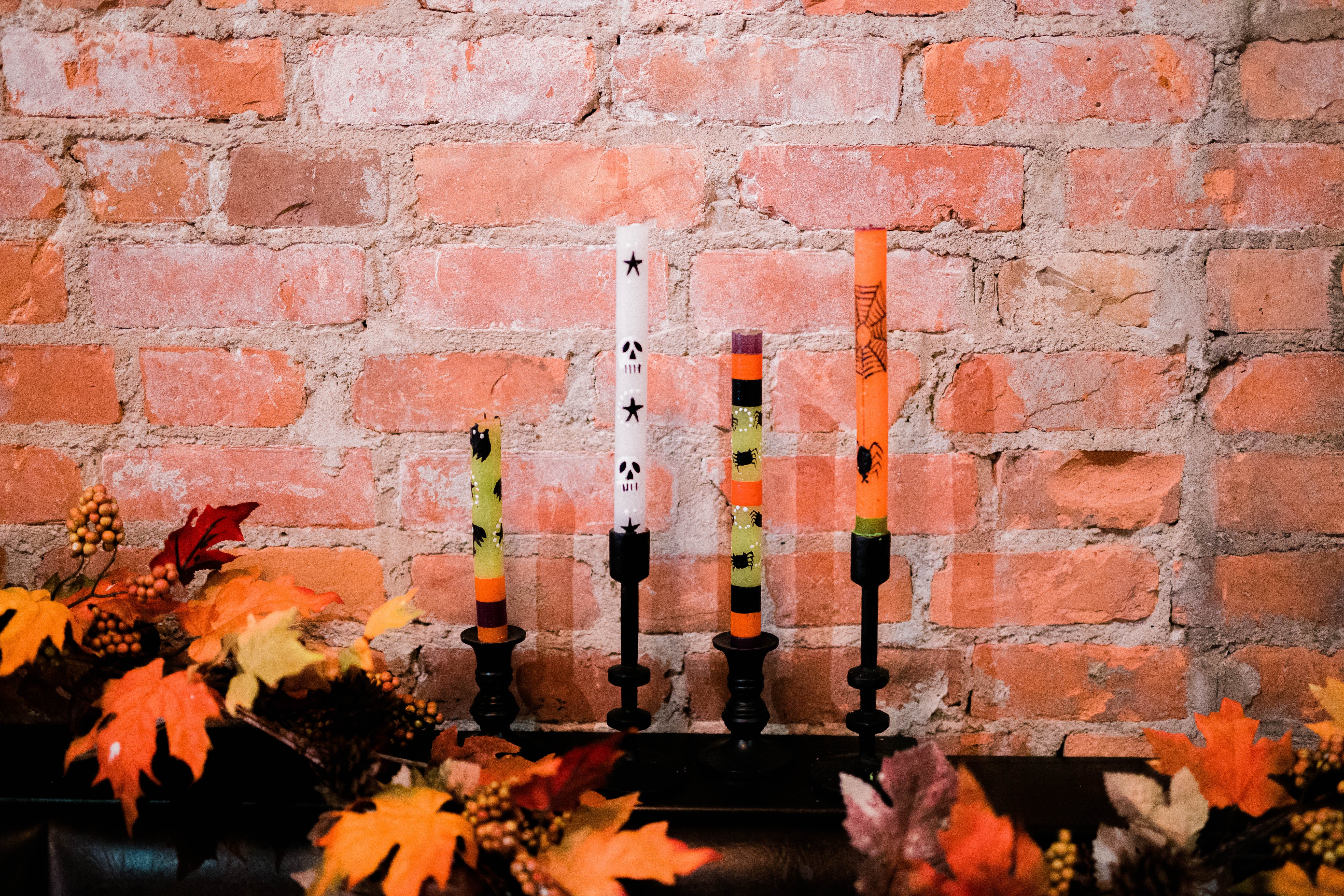 Four Halloween hand made and hand painted taper candles in black taper candle holders with autumn leaves in the foreground and brick wall in the background for a 'lifestyle' shot of the Halloween candles.