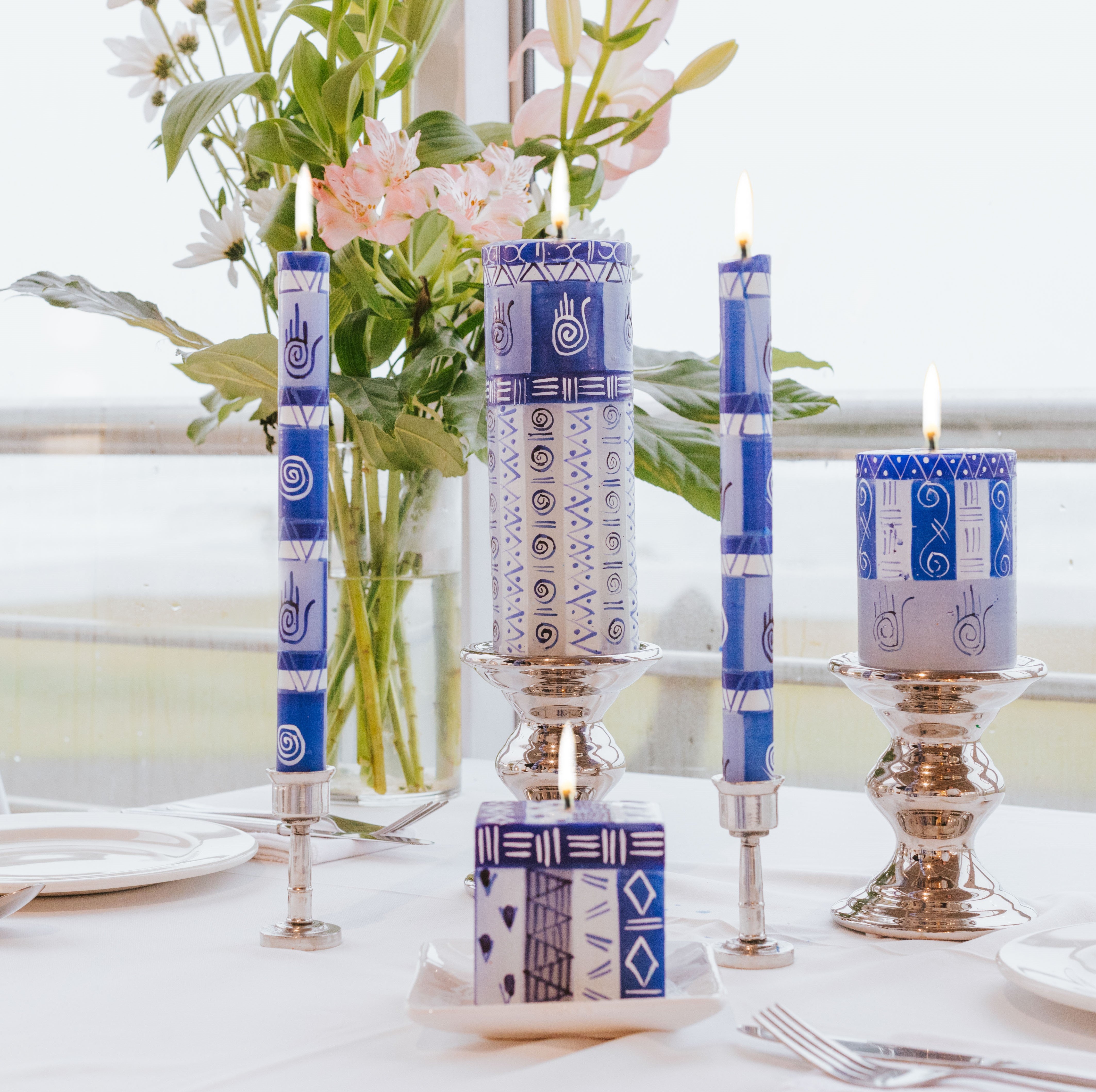 Lifestyle photo of hand painted Hamsa candles in Blue & White and include the Hamsa Hand to protect from evil spirits! Table setting with taper pair in pewter taper holders, 3' x 4' pillar, 3" x 6 " pillar, in silver holders, and 3' x 3' x 3' cube candle in candle coaster, all burning brightly as the center piece.