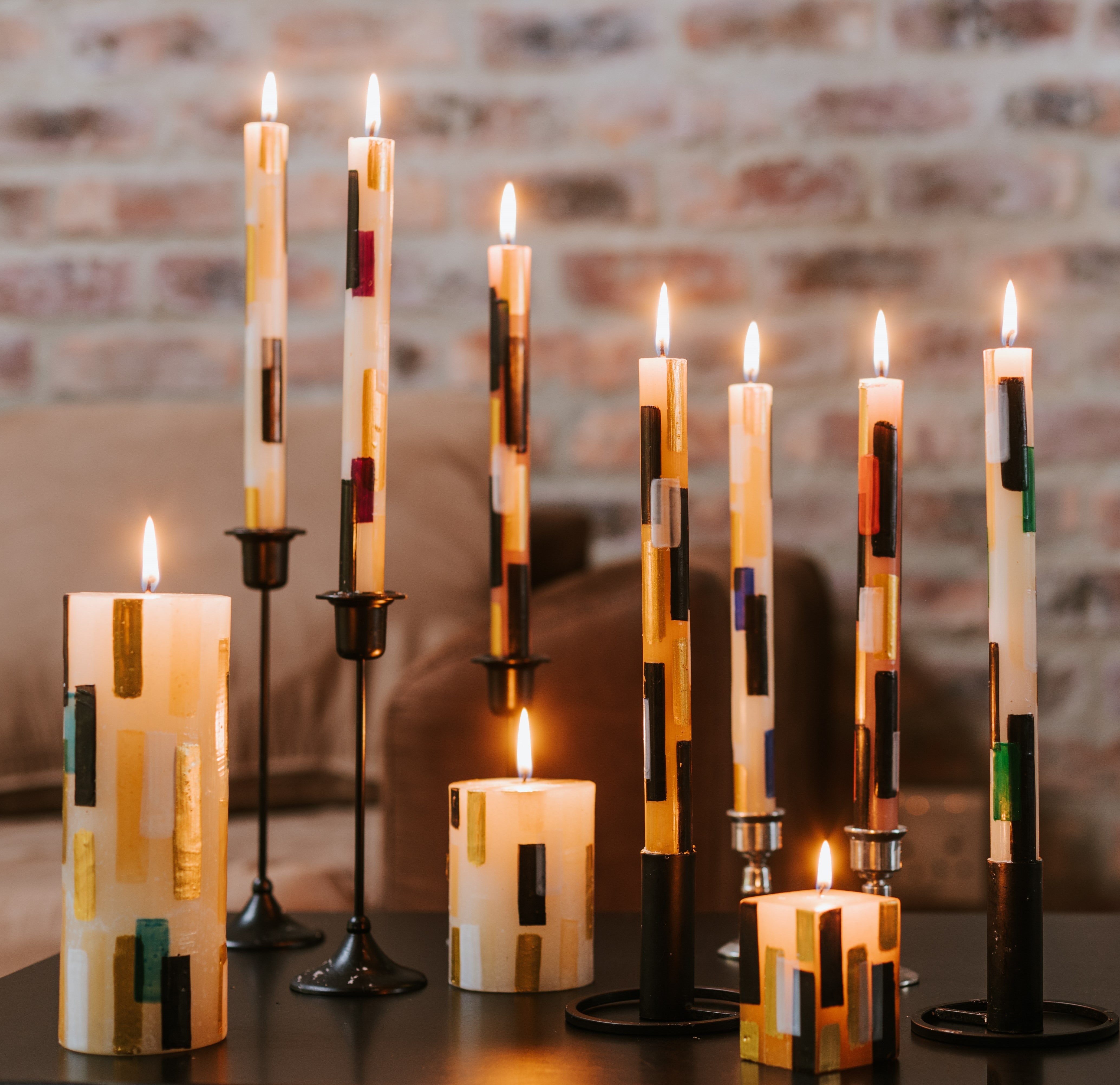 Lifestyle photo of the Klimt collection; tapers 3x8 pillar, 3x4 pillar, and 2x2x3 cube. Tapers are all different heights and all candles are lit with a soft brick wall in the background - just beautiful!
