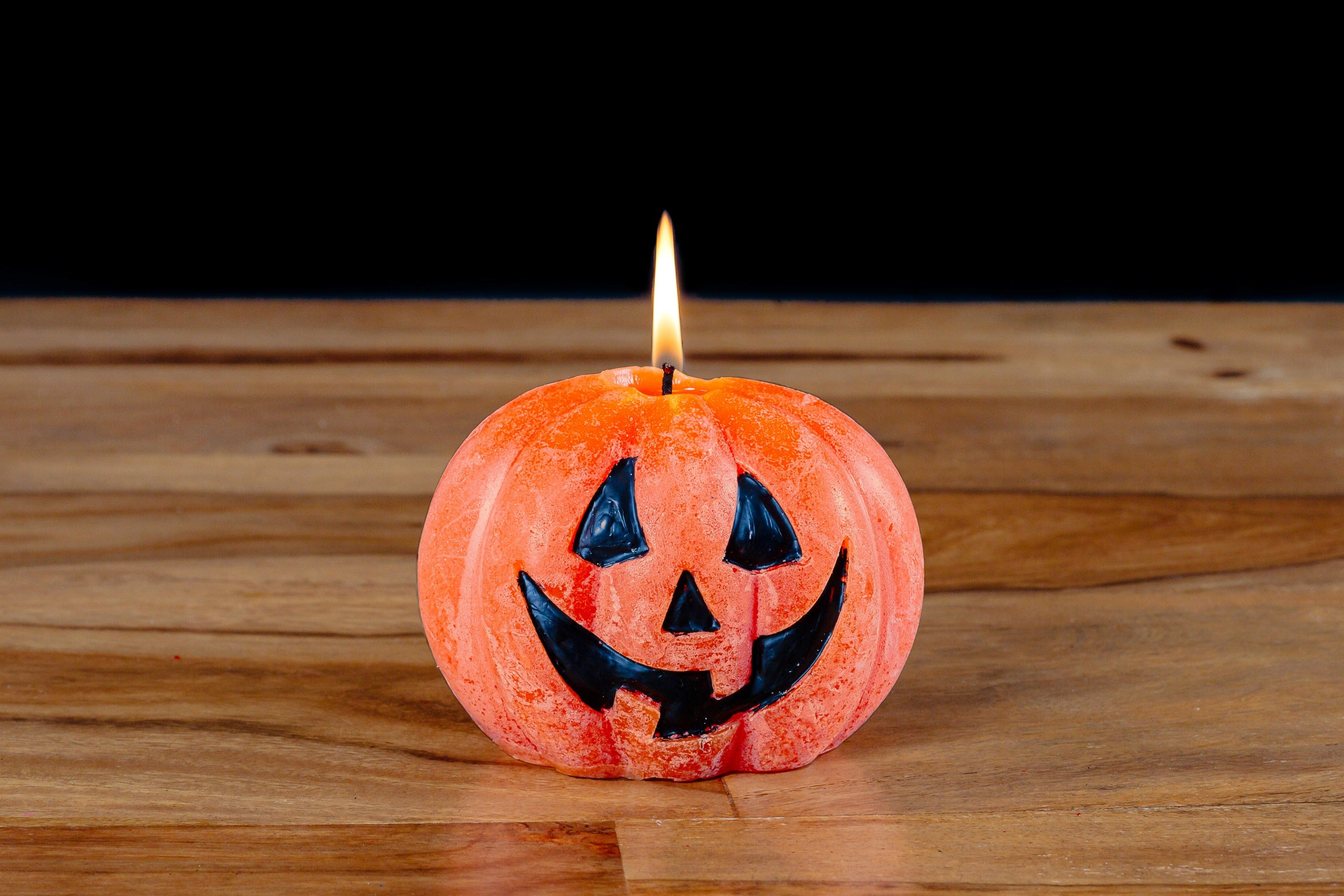 Whimsy Pumpkin candle! Orange with a Jack o' Lantern face painted on in black. This pumpkin is lit sitting on a wooden table with a black background - BOO!