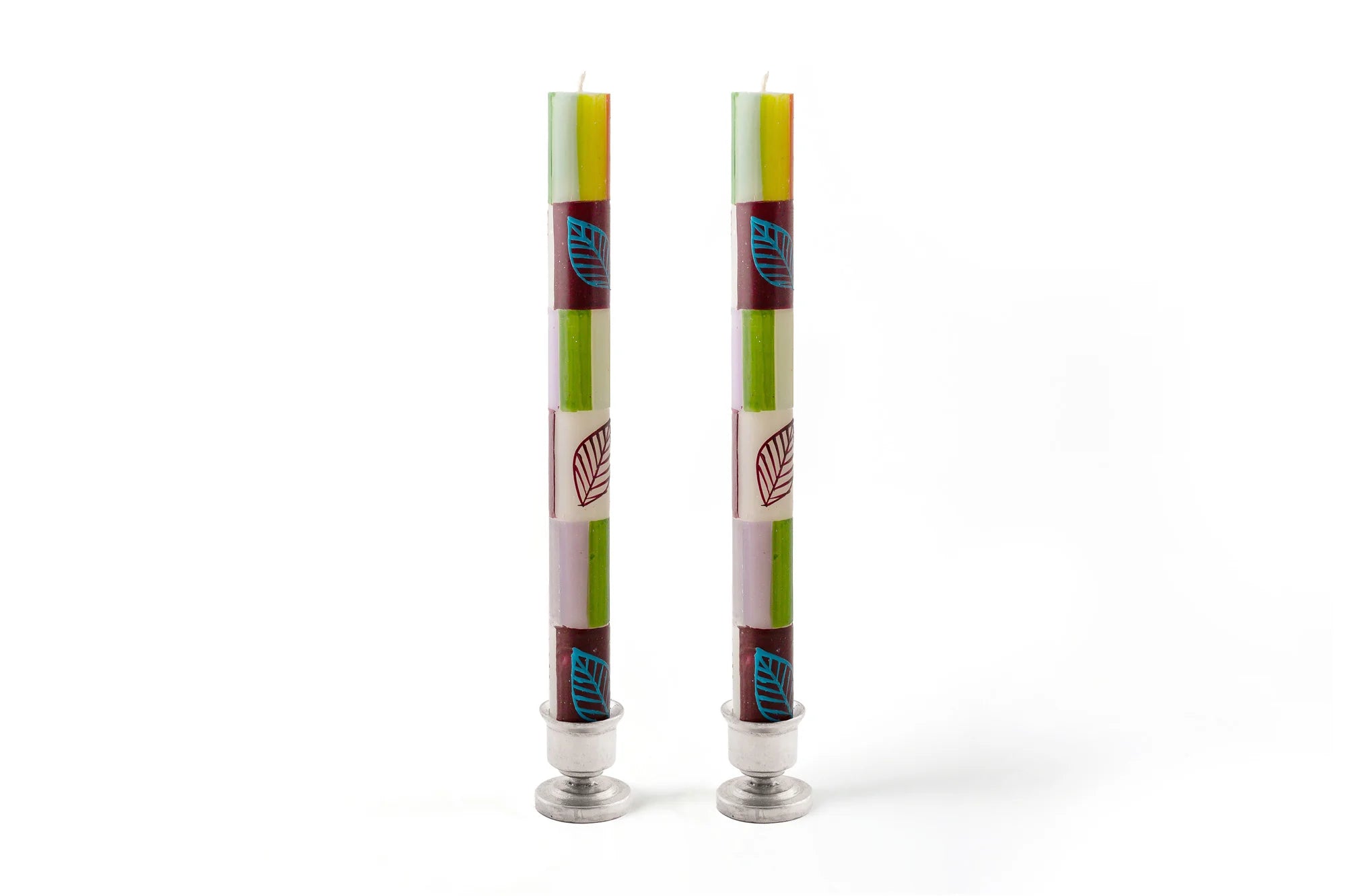 Pair of matched Magic Garden taper candles n small pewter taper candle holders. The tapers are a white base with green, orange, turquoise, and lavender stripes. Overlay of pigment colors with a variety floral and leaf designs.