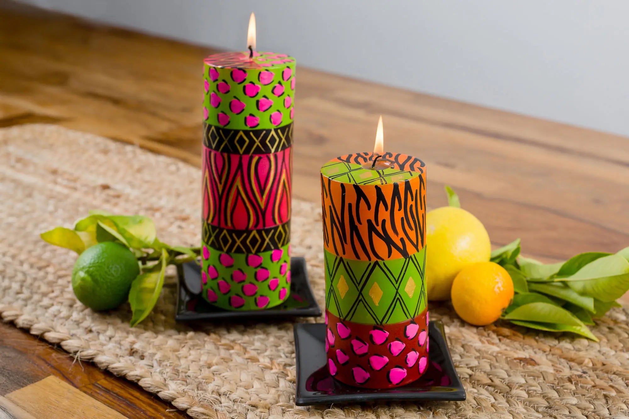 Lifestyle photo of the Wild Animal Print 3" x 6" pillar candle and 3" x 8" pillar candle, on black candle coasters. Each burning brightly on a jute mate on a wooden table.   