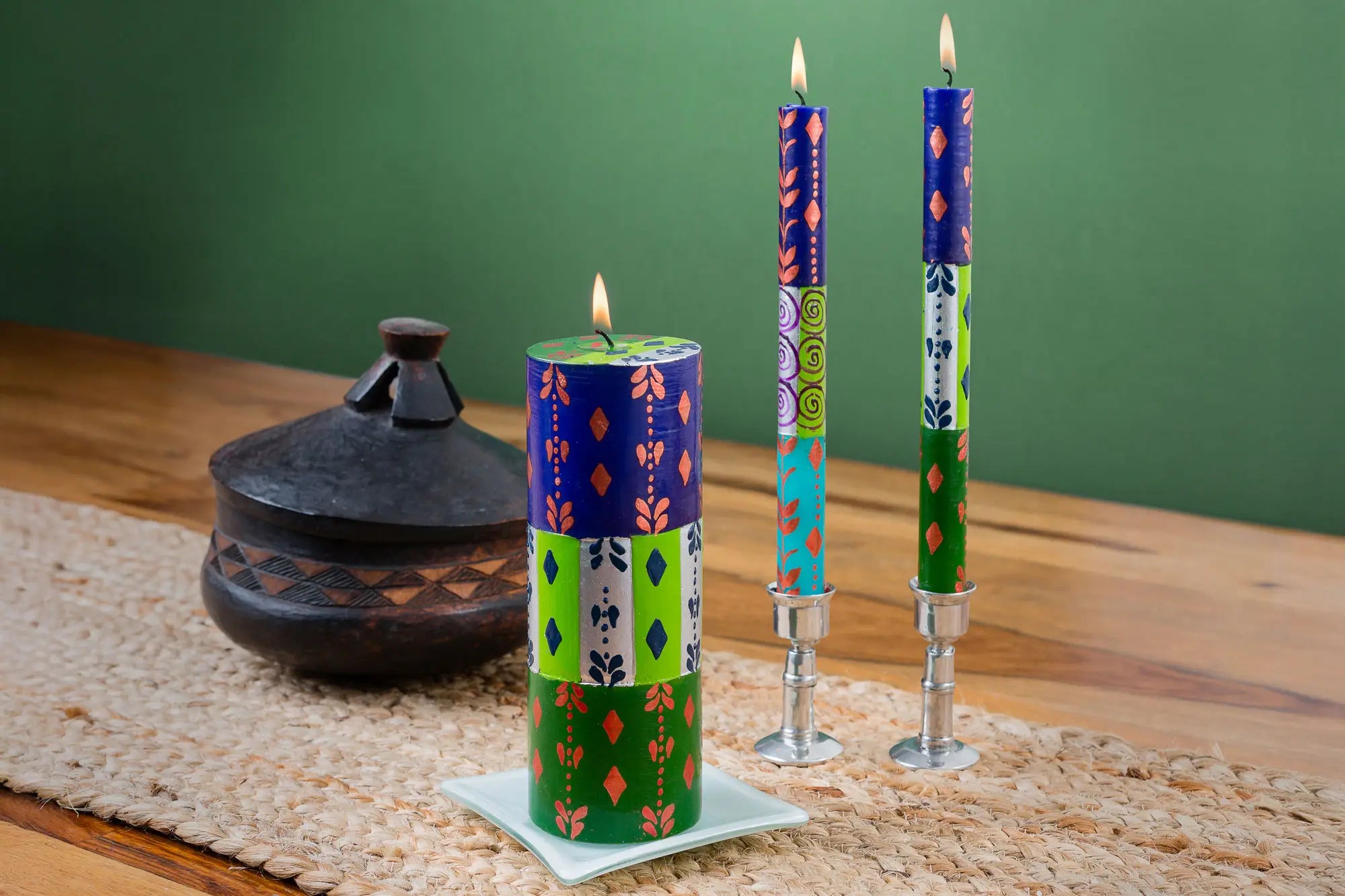 Life-style photo of the Sari 3" x 8" pillar candle on a white candle coaster, alongside a matched pair of Sari tapers in pewter taper candle holders.  All burning on a table.  Beautiful!