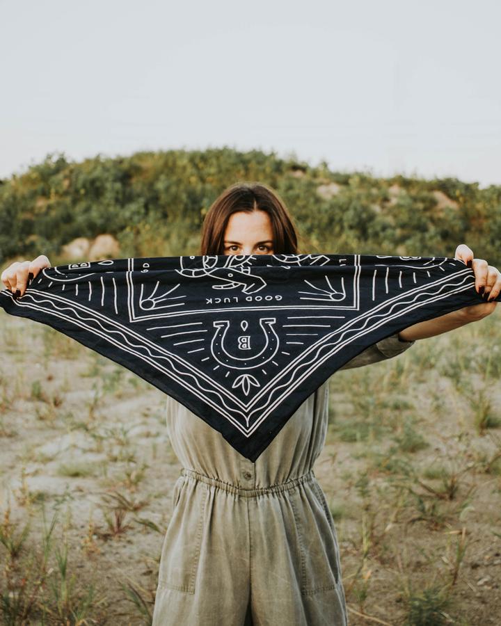 Woman streching the Good Luck bandana out to show the design of horse shoes and the message 'Good Luck'  Dark navy blue with cream color outline design.