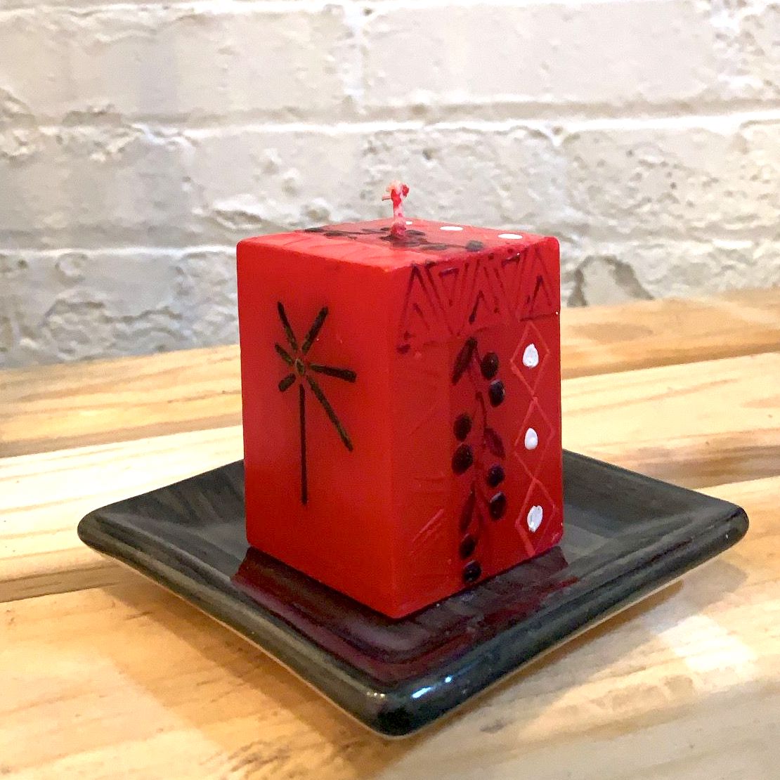 Kwanzaa red 2x2x3 cube candle showing symbol on the front and flower pattern on the side. Sitting on a black candle coaster.