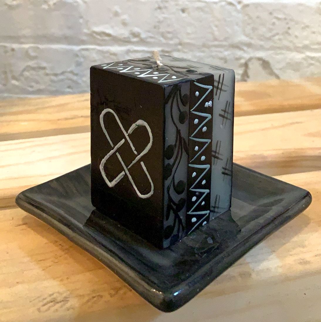 Kwanzaa black 2x2x3 cube candle showing the symbol on the front and the flower design on the side.  Sitting on a black candle coaster.  Fair Trade.