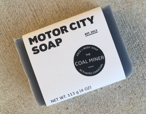 Coal Miner soap bar that is dark grey with Motor City branded paper wrap that is  cream color with MCS logo and label at Coal Miner - charcoal soap! 4oz bar.