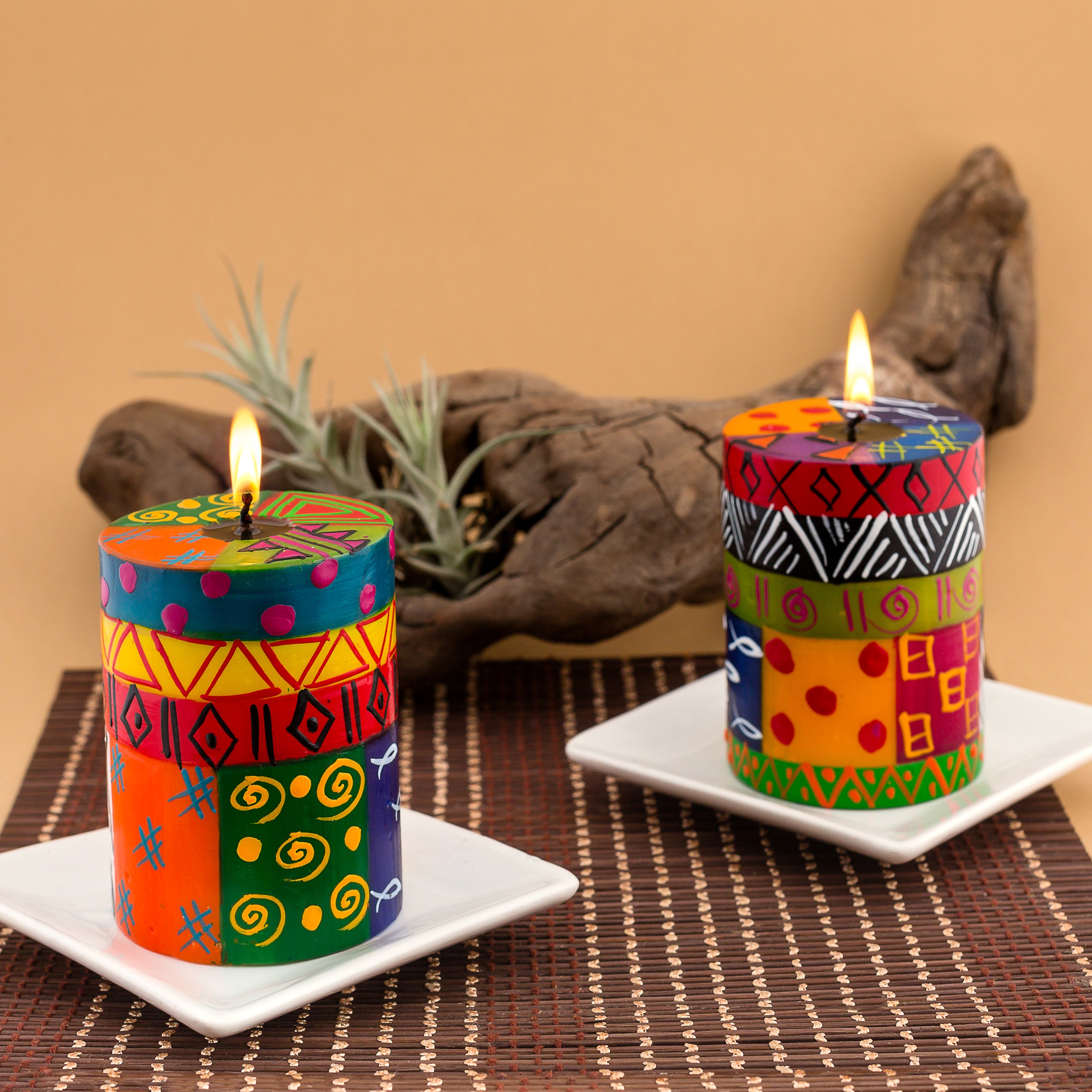 Two Multicolor Ethnic 3x4 pillar candles on white candle coasters. Both candles are lite, and sitting on a woven placemat with a plant behind them.  Lovely lifestyle shot of what they look like in a home.