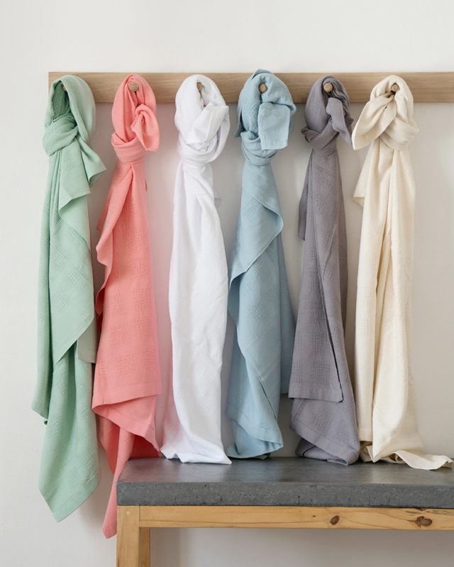 Organic cotton baby blankets hanging on a coat rack. Tied at the top and hanging down, in the colors of light green, light orange, white, light turquoise, elephant grey, and cream.