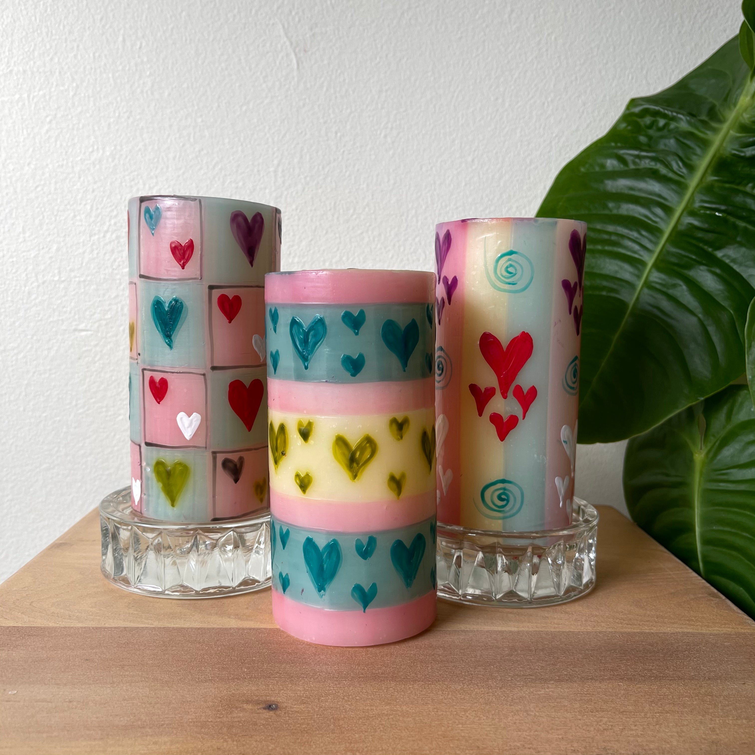 Three Pastel Heart 3x6 pillar candles.  Two are sitting on glass candle coasters.  All painted with hearts of various size & colors on pastel background. Very fun!