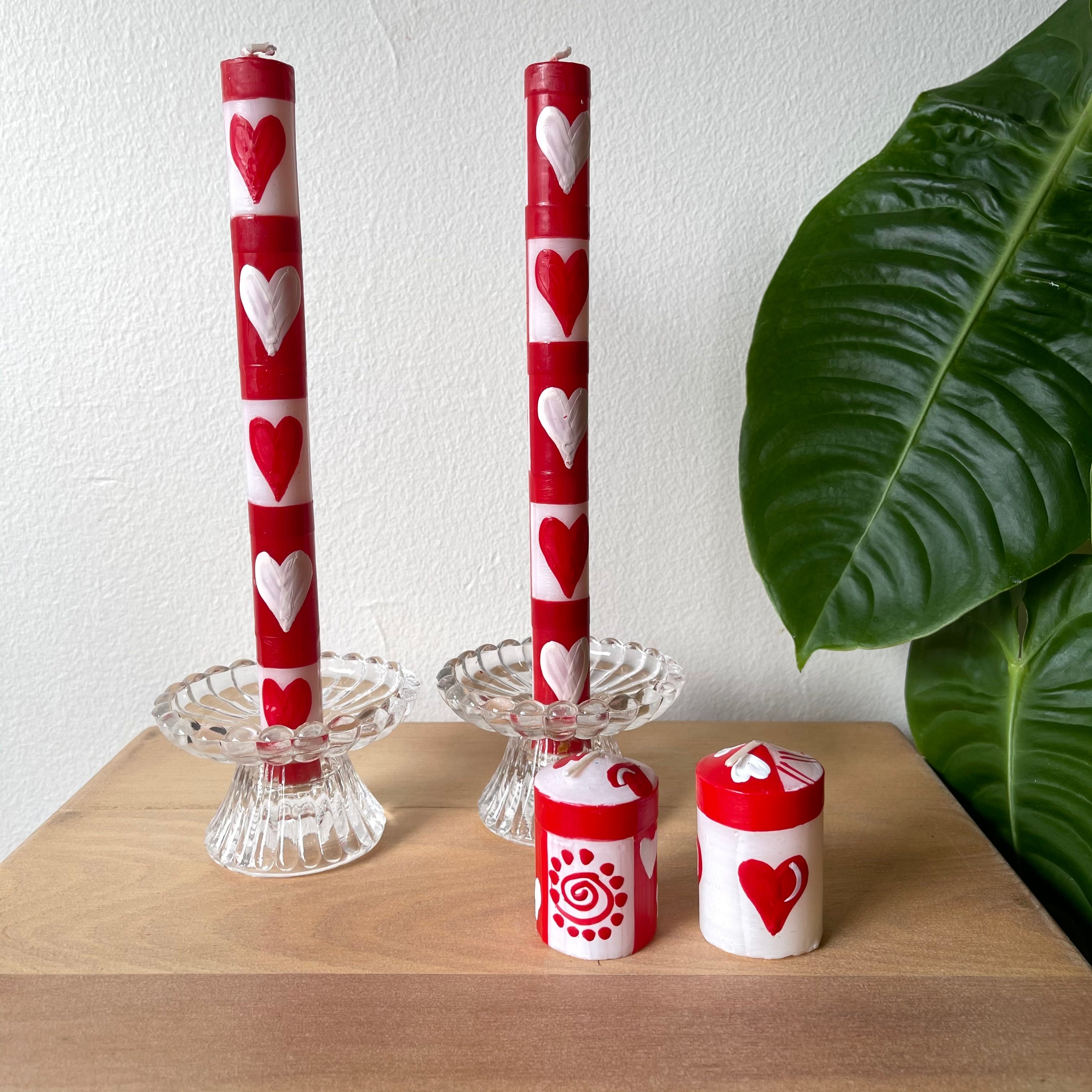 A pair of Valentines taper candles in glass taper holders, with a pair of votive candles in front of them in more of a life-style setting with a plant on the side.  The candles are hand painted with red & white hearts!