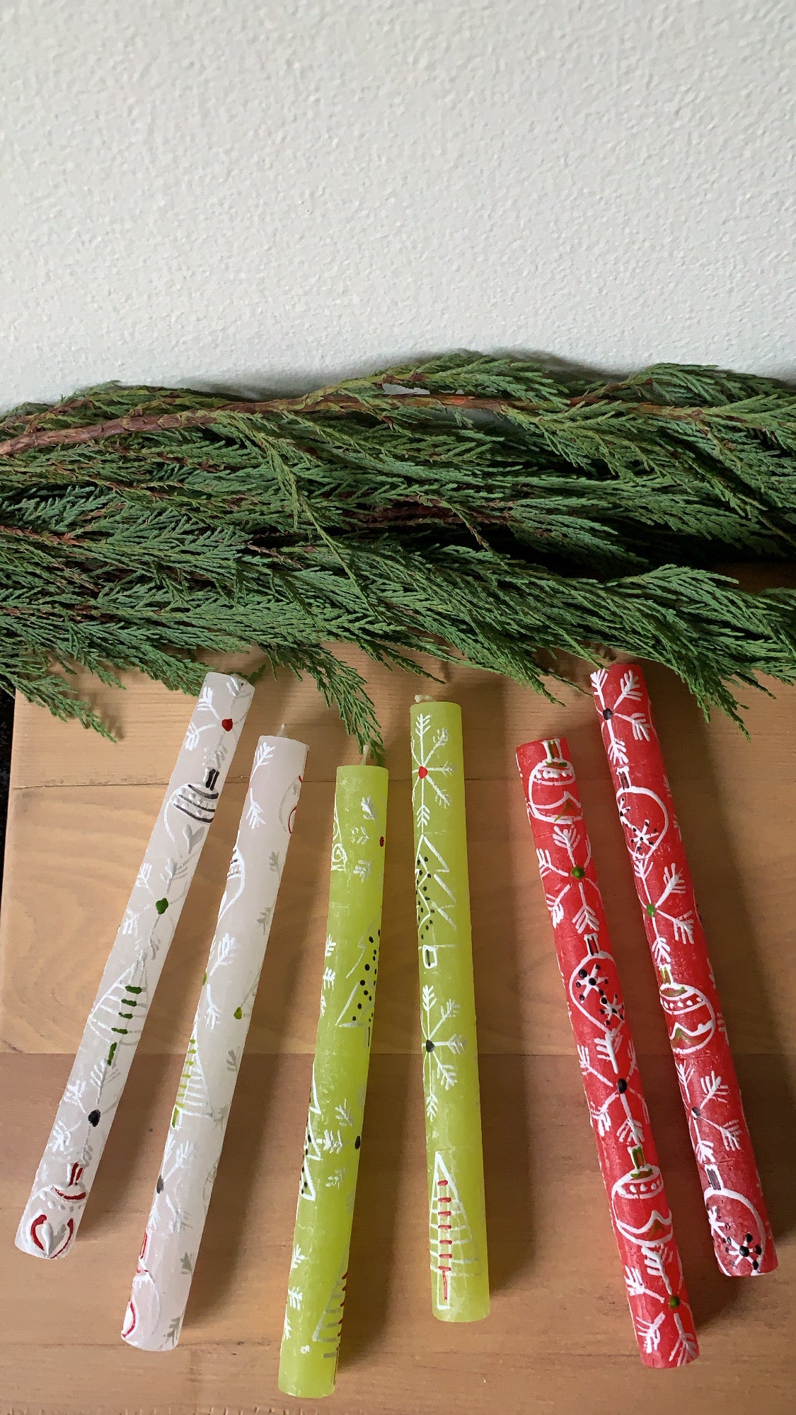Three pairs of Whimsy Christmas taper candles; base candles are red, white & green, each hand painted with 'whimsy' Christmas designs such as snow flakes, trees, and ornaments in white with accent colors of red and green. These are presented on a table with green fur tree.